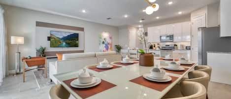 A modern dining area with a long table set for six, a sofa, and a kitchen in the background.