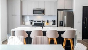 A modern kitchen with white cabinetry, a stainless steel refrigerator, a microwave, and a coffee machine. There are four bar stools and a white dining table.
