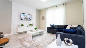 A modern living room with a flat-screen TV mounted on a textured wall, a grey sofa with blue cushions, a white coffee table, and a decorative vase. - 4BR Home with Private Pool & Disney Themes (DAVENPORT)