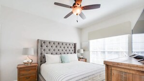 A bedroom with a tufted headboard, a bed with striped bedding, two bedside tables with lamps, a wooden desk, and a ceiling fan.