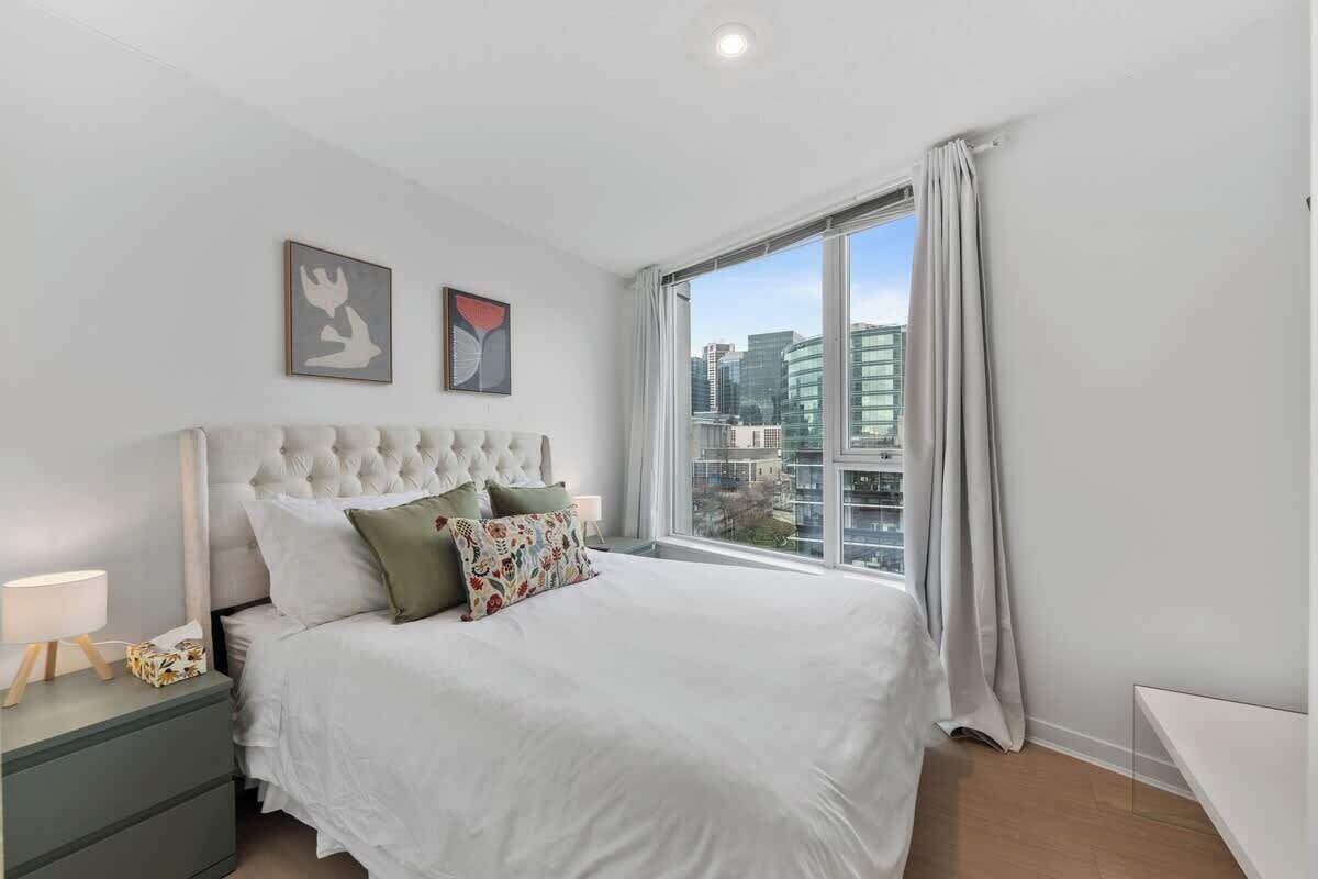 A bedroom with a large bed, a nightstand, a window with curtains, and two wall-mounted artworks.