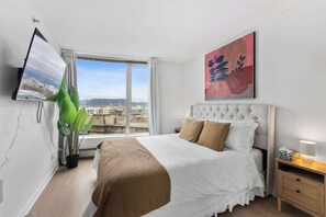 Habitación de hotel con cama, televisor montado en la pared, ventana con vistas a edificios y una planta en maceta.