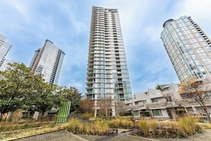 A tall residential building with multiple balconies, surrounded by greenery and other buildings.