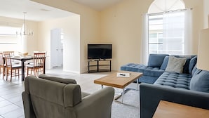 A modern living room with a sofa, a coffee table, a TV, and a dining area with chairs and a table. - Beautiful Retreat *5min -> Disney *Pool *King Bed (Kissimmee)