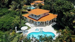 Une maison à deux étages avec une piscine, le tout entouré d’une végétation luxuriante.