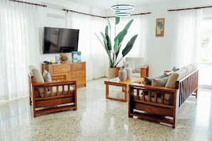 Een woonkamer met een televisie, houten meubels, een grote plant en een raam met gordijnen.