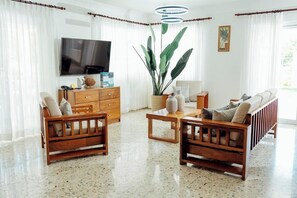 A living room with a TV, wooden furniture, a large plant, and a window with curtains.