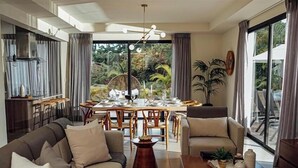 Un salon moderne comprenant un canapé, un fauteuil, une table à manger et des chaises. On y trouve une grande fenêtre avec des rideaux, un luminaire au plafond et une plante sur la table.