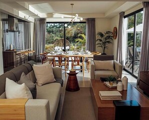 Ein modernes Wohnzimmer mit Sofa, Sesseln, Esstisch und Stühlen. Ein großes Fenster mit Vorhängen, eine Deckenleuchte und eine Pflanze auf dem Tisch.