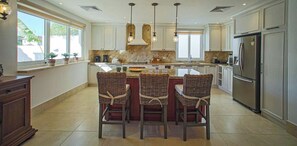 A spacious kitchen with a central island, three wicker chairs, and a large refrigerator.