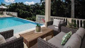 Uma área junto à piscina com mobiliário de vime e uma mesa de madeira, rodeada por vegetação exuberante.
