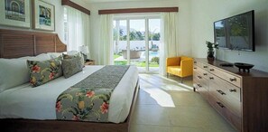 A hotel room with a large bed, a wooden headboard, a television, a desk with drawers, a chair, and a view of a pool and greenery outside.