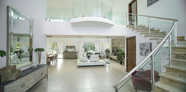A spacious living area with a high ceiling, a staircase, and a dining area in the background.