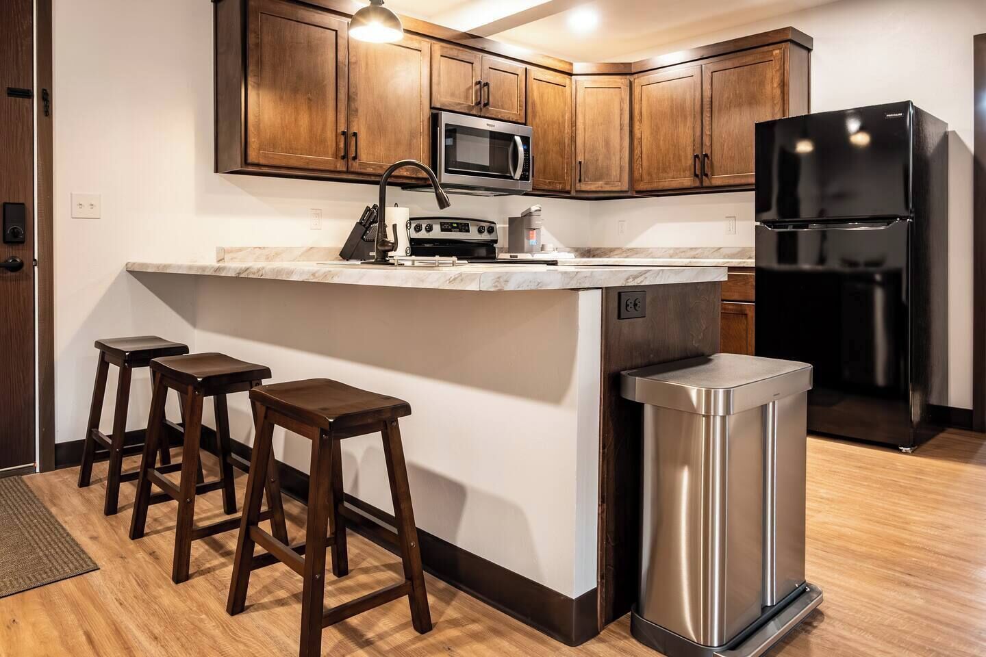A modern kitchen with wooden cabinets, a stainless steel refrigerator, a microwave, and a sink. There are three wooden stools at the counter.