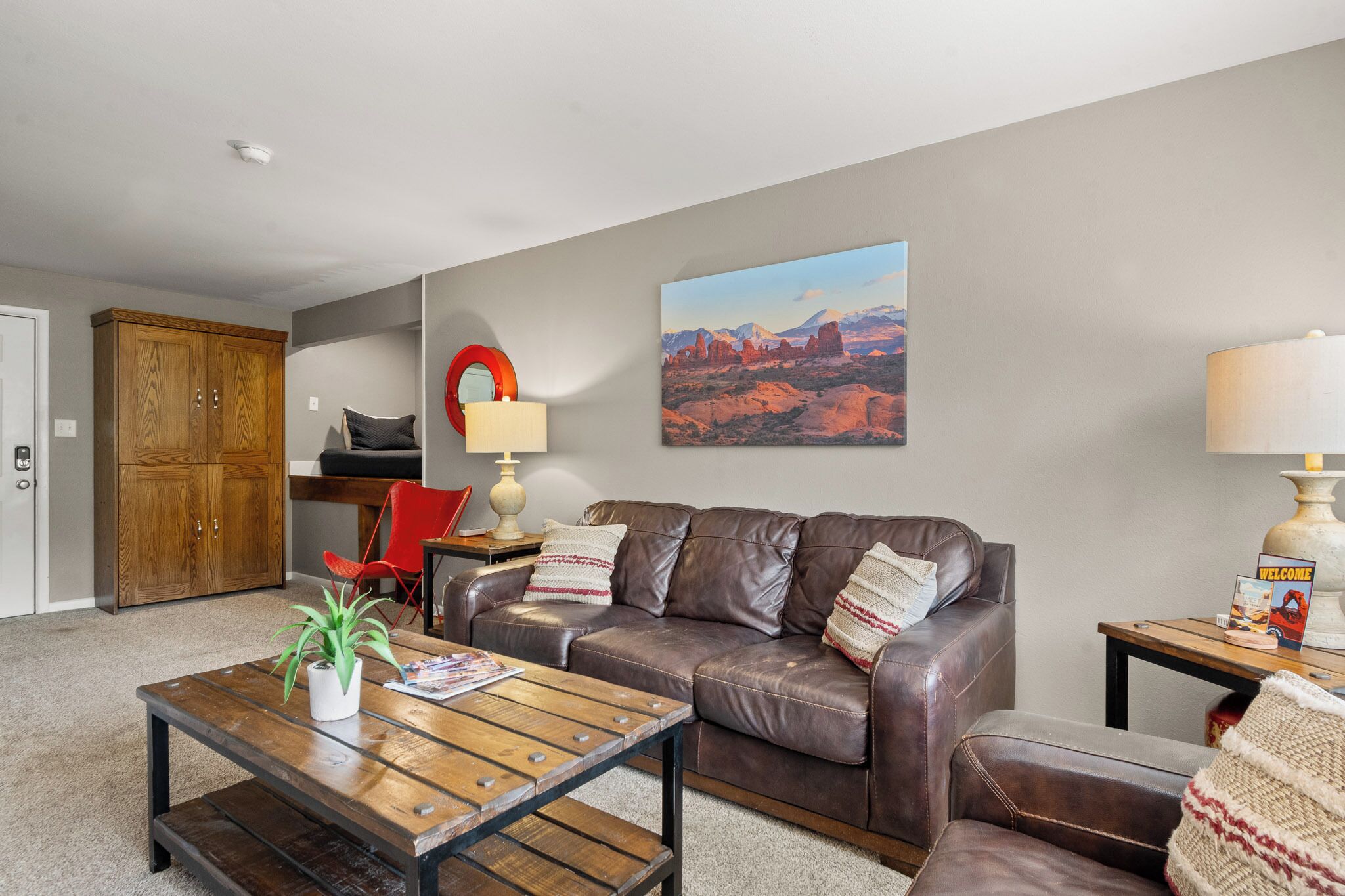 A living room with a brown leather couch, a wooden coffee table, a red chair, a lamp, and a painting of a desert landscape.