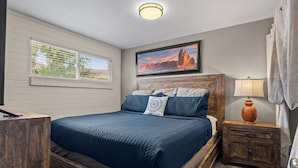 Een slaapkamer met een groot bed, een houten hoofdbord, een nachtkastje met een lamp, een raam met jaloezieën en een lijst met een afbeelding aan de muur.
