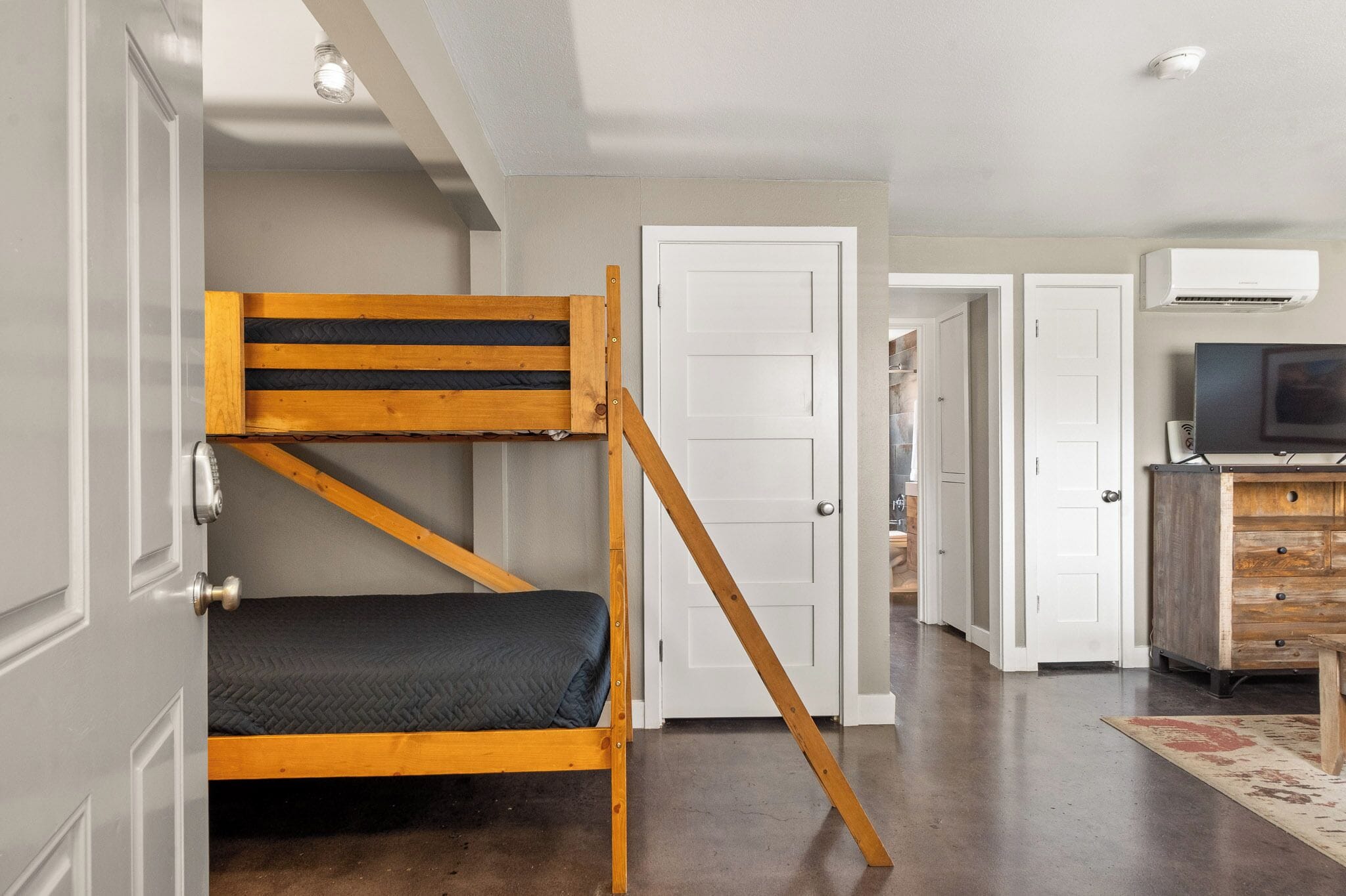 Un lit superposé avec une structure en bois et un matelas noir, une porte blanche, une commode blanche avec des tiroirs et un téléviseur posé sur le dessus.