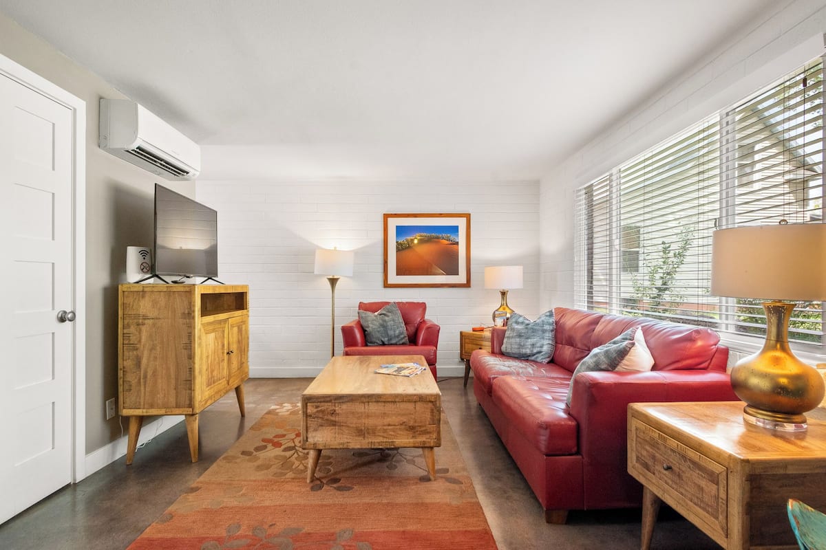 Una sala de estar con un sofá rojo, una mesa de centro de madera y un televisor sobre un mueble de madera. Hay una ventana con persianas y un cuadro en la pared.