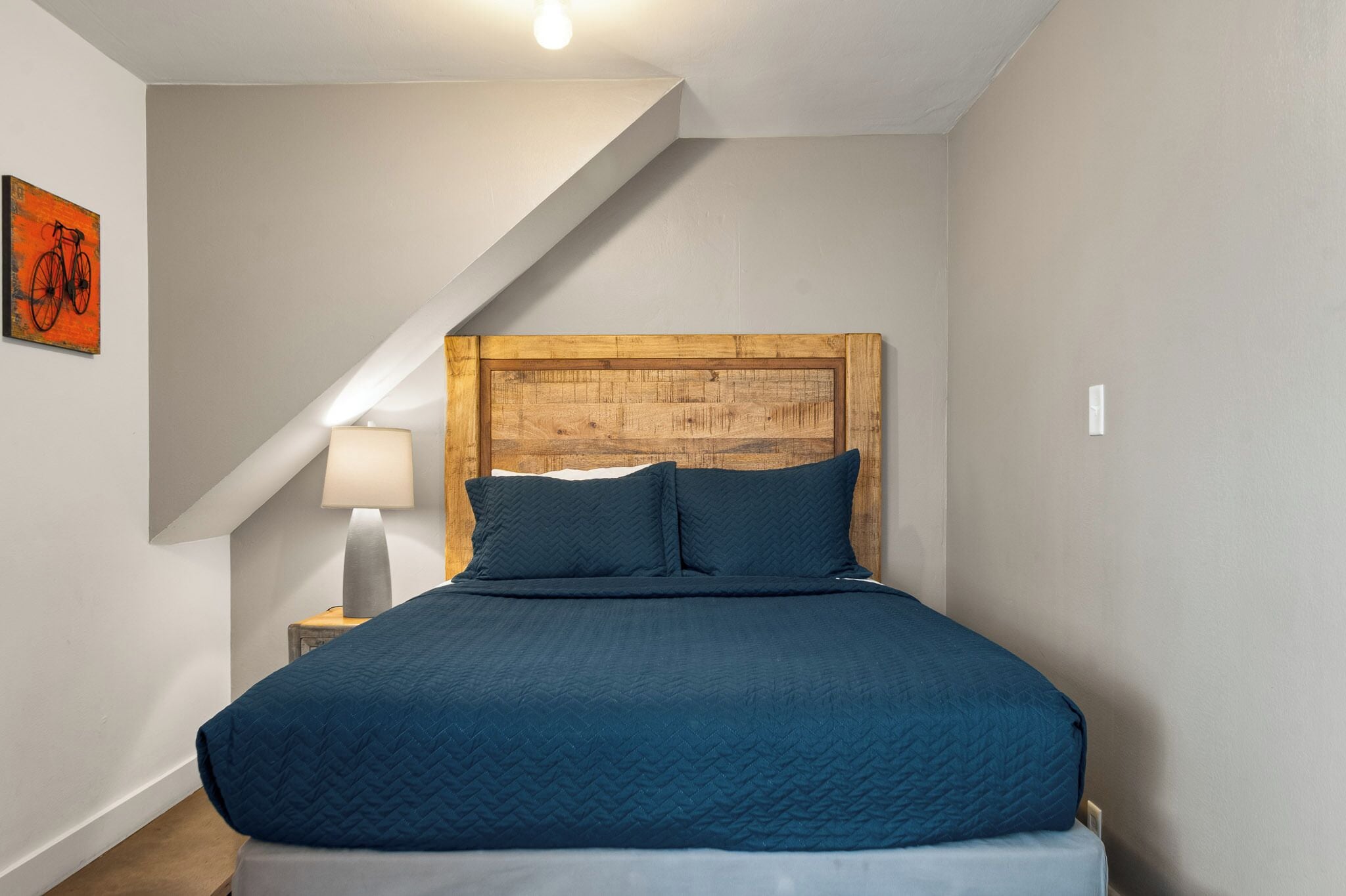 A bedroom with a wooden headboard, a blue quilted bedspread, a bedside lamp, and a wall-mounted bicycle artwork.
