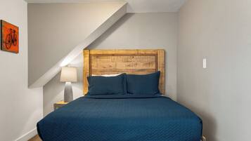 A bedroom with a wooden headboard, a blue quilted bedspread, a bedside lamp, and a wall-mounted bicycle artwork.