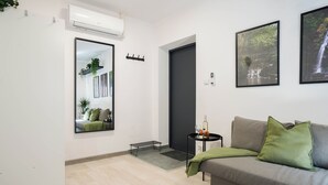 Una sala de estar moderna con un sofá gris, cojines verdes, una mesita auxiliar con una botella y vasos, otra mesita auxiliar negra, un espejo y dos cuadros enmarcados en la pared.
