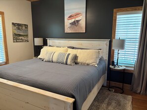 A bedroom with a bed, a nightstand, a lamp, a window with blinds, and wall art. - Lakefront Home, New Dock, Stone Fire Pit Overlooking Lake Wedowee, Sleeps 10 (Woodland)
