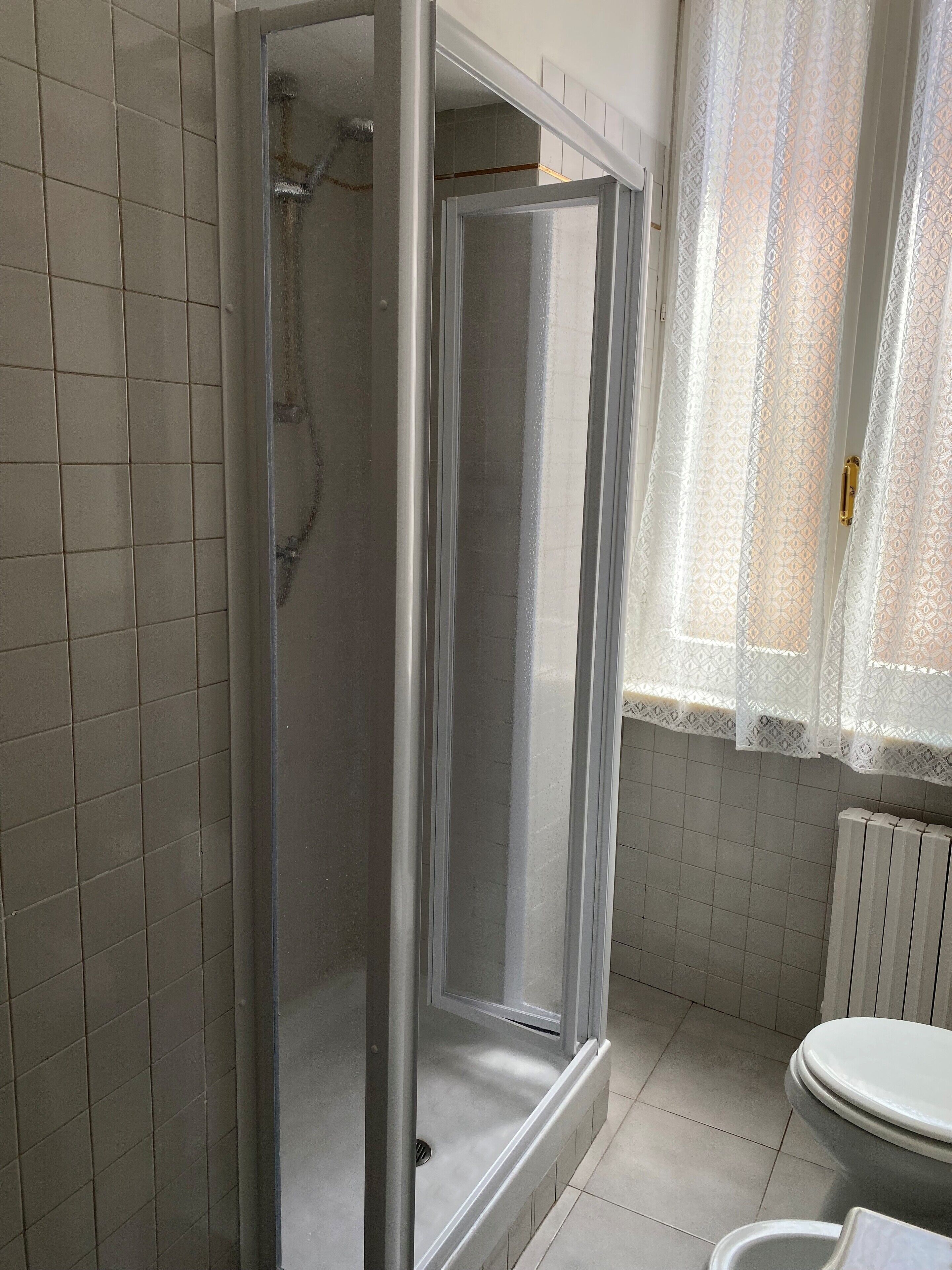 A bathroom with a glass-enclosed shower, tiled walls, a toilet, and a window with patterned curtains.