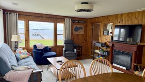 A cozy living room with a sofa, a dining table, and a fireplace. Large windows offer a view of a lake and dock.