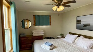 A bedroom with a bed, a bedside table, a dresser, a ceiling fan, a window with curtains, a mirror, and a wall-mounted painting.