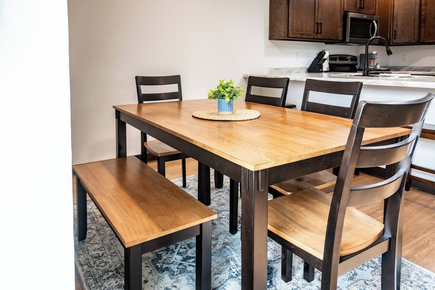 Un comedor con una mesa y sillas de madera, una planta pequeña en la mesa y una cocina al fondo.