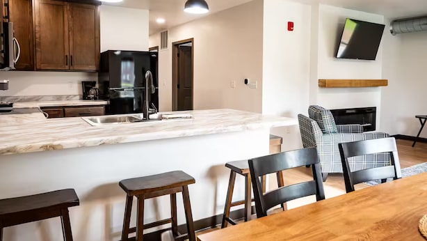 A modern kitchen with a marble countertop, wooden cabinets, and a built-in stove. There is a dining area with a wooden table and chairs, and a living room with a sofa and a flat-screen TV mounted on the wall. - Brillion 2‑Bedroom Suite w/ Full Kitchen Near Ariens – Unit #4 (Brillion)