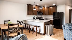 A modern kitchen with wooden cabinets, a central island, a black refrigerator, and a dining area with a table and chairs. - Brillion 2‑Bedroom Suite w/ Full Kitchen Near Ariens – Unit #4 (Brillion)