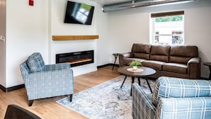 A modern living room with a fireplace, a sofa, a TV, and a window with a view of greenery. - Brillion 2‑Bedroom Suite w/ Full Kitchen Near Ariens – Unit #4 (Brillion)