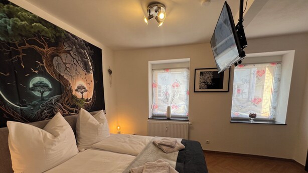 A bedroom with a bed, a flat-screen TV mounted on the wall, two windows with floral curtains, and a tapestry on the wall. - ❤ VipStay Stylish 57m² ground-floor app., kitchen, terrace, Wi-Fi. Near S-Bahn ❤ (Eschelbronn)