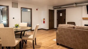 A modern living room with a wooden floor, a round dining table with chairs, a sofa, and a fire extinguisher on the wall. - Brillion 2‑Bedroom Suite w/ Full Kitchen Near Ariens – Unit #5 (Brillion)