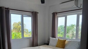Une chambre à coucher avec un lit, un ventilateur de plafond, un climatiseur et de grandes fenêtres avec des rideaux.