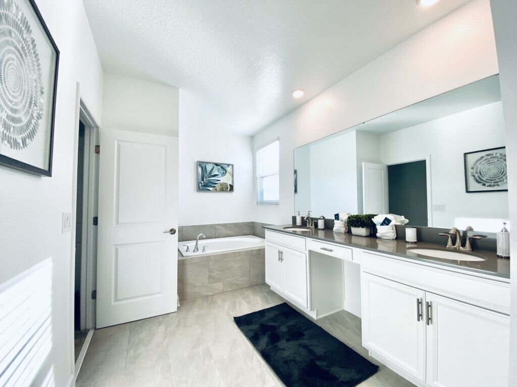 A modern bathroom with a large mirror, two sinks, a bathtub, and a door leading to another room.