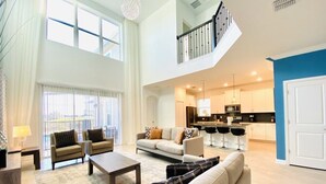A spacious living area with a high ceiling, a kitchen in the background, and a staircase leading to an upper level.