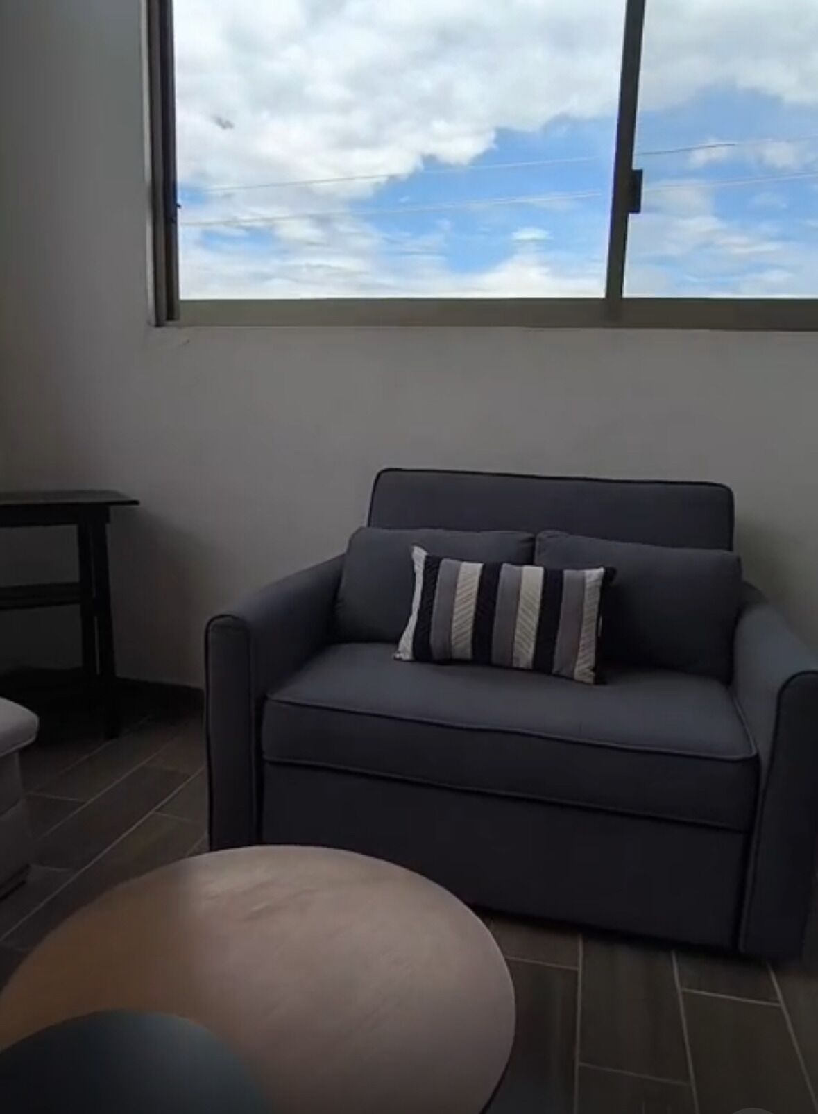 Una sala de estar moderna con un sofá gris, una pequeña mesa redonda y vistas del cielo a través de un amplio ventanal.