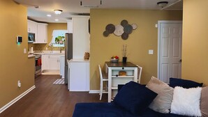 Un salon moderne doté d’un ventilateur de plafond, un coin repas avec une table et des chaises, une cuisine avec des meubles blancs et une porte donnant sur une autre pièce.