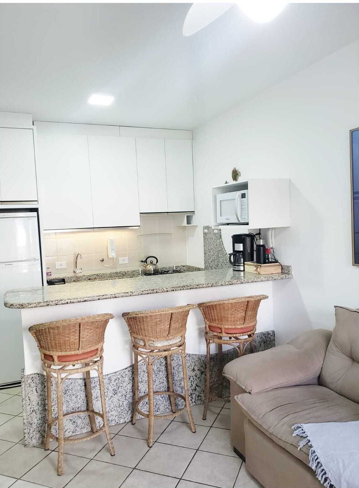 A compact kitchen with white cabinets, a granite countertop, a microwave, and a coffee maker. There are three wicker bar stools and a beige sofa.