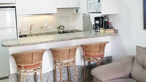 A compact kitchen with white cabinets, a granite countertop, a microwave, and a coffee maker. There are three wicker bar stools and a beige sofa. - Apartment in Ingleses Norte Floriañópolis (Florianópolis)