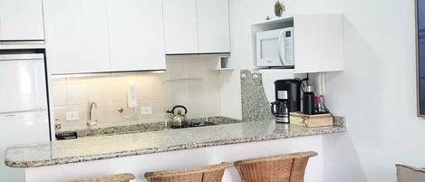 A compact kitchen with white cabinets, a granite countertop, a microwave, and a coffee maker. There are three wicker bar stools and a beige sofa.