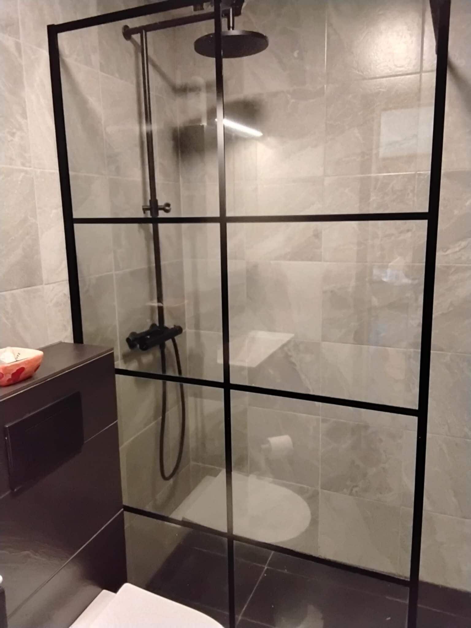 Un espace douche clos par des parois de verre, avec un cadre noir, des murs carrelés et un sol foncé.