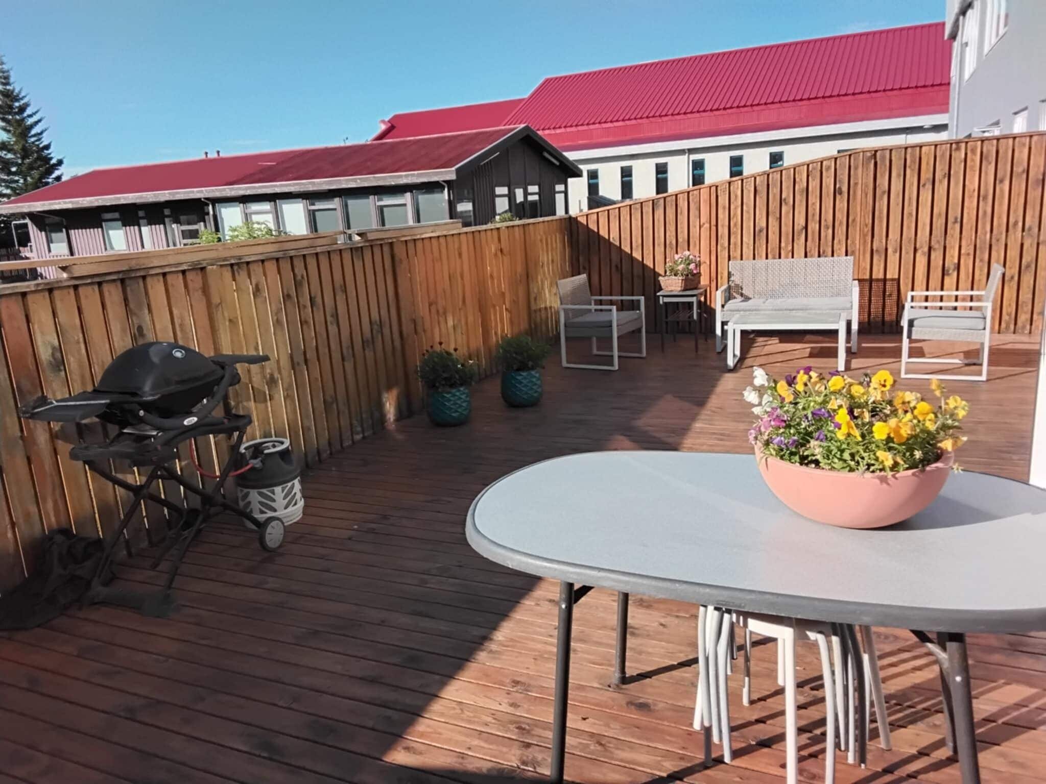 En træterrasse med grill, et rundt bord og en blomsterpotte, samt udendørs møbler.