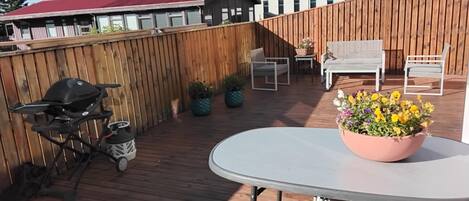 En træterrasse med grill, et rundt bord og en blomsterpotte, samt udendørs møbler.
