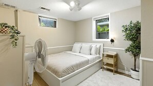 Ein Schlafzimmer mit einem Bett, einem Standventilator, einem Nachttisch, einer Wandlampe und einer Topfpflanze.