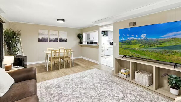 Ein Wohnzimmer mit braunem Ledersofa, einem Esstisch mit Stühlen, einem großen Flachbildfernseher auf einem Standfuß, einem gemusterten Teppich und einer Küche im Hintergrund.