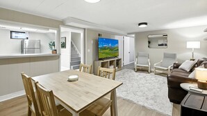 Ein modernes Wohnzimmer mit einem Holzesstisch, zwei Stühlen, einer Couch, einem Fernseher und einer Küchenzeile inklusive Kühlschrank und einer Pflanze.