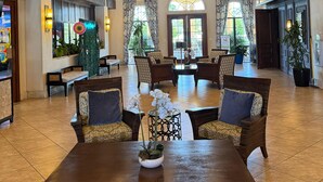 Eine geräumige Lobby mit hohen Decken, Holzbalken und mehreren Deckenventilatoren. Es gibt verschiedene Sitzbereiche mit Stühlen und Tischen, und eine große Zierpflanze in der Mitte.